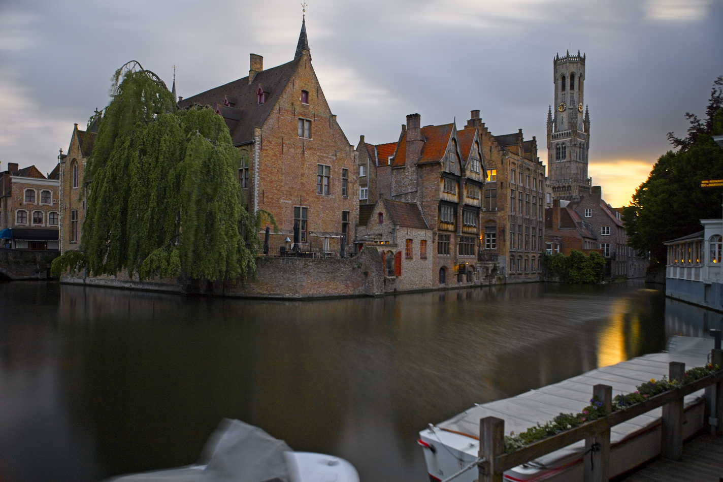 A picturesque view across a dark canal toward historic European architecture at twilight, featuring a cluster of old buildings, a prominent belfry tower, and a large weeping tree framing the scene under a dramatic sky.